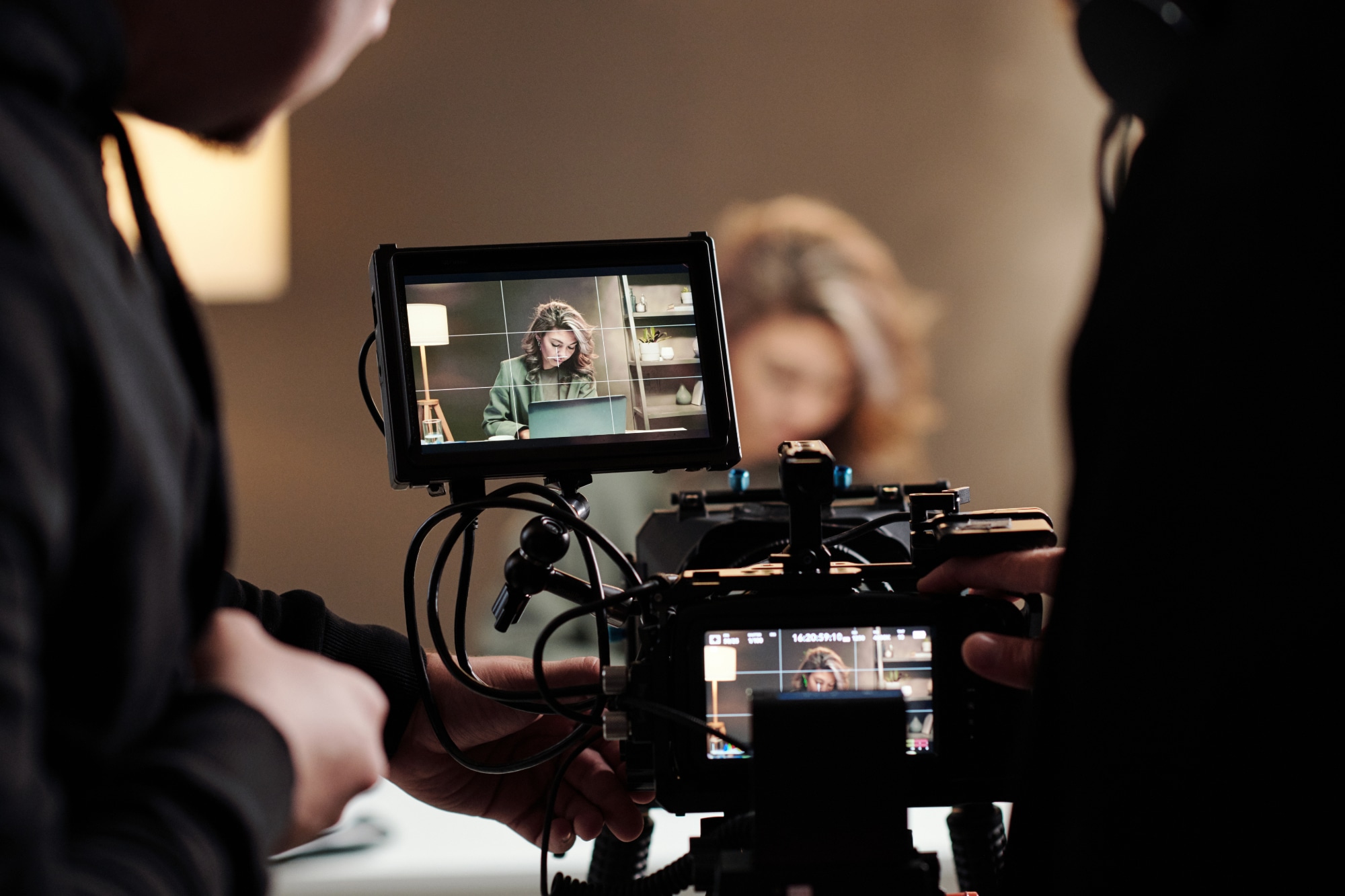 A behind-the-scenes view of a film shoot, showing a steadicam monitor with a female actor working on a laptop at a table, while a cameraman and assistant frame the shot.</p>
<p>